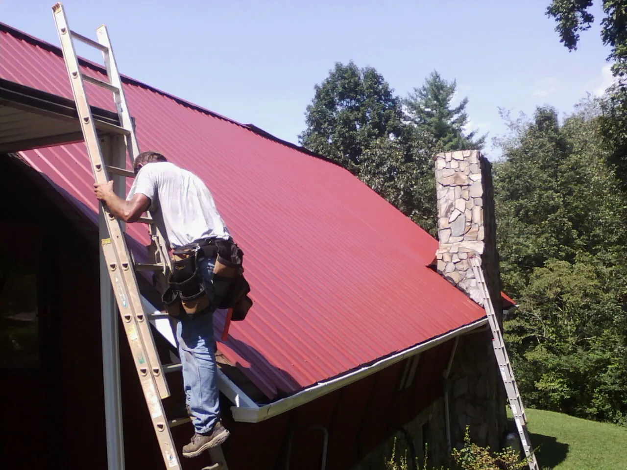 Roofer on a ladder beside a red metal roof and stone chimney — Soffit & Fascia Repair by Roofing Brothers in Bethpage